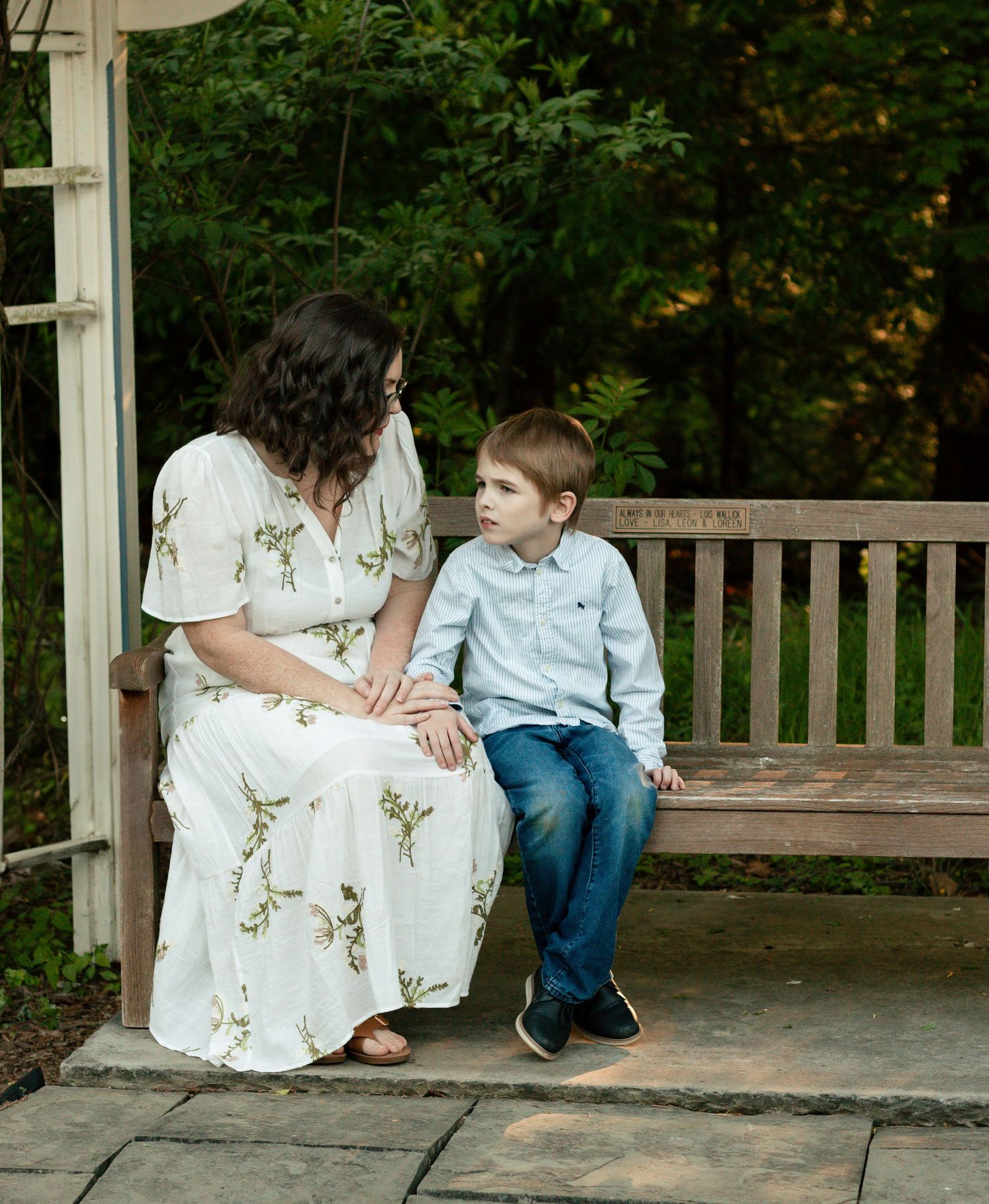 Two people share a quiet moment together while sitting on a wooden bench in a garden setting.