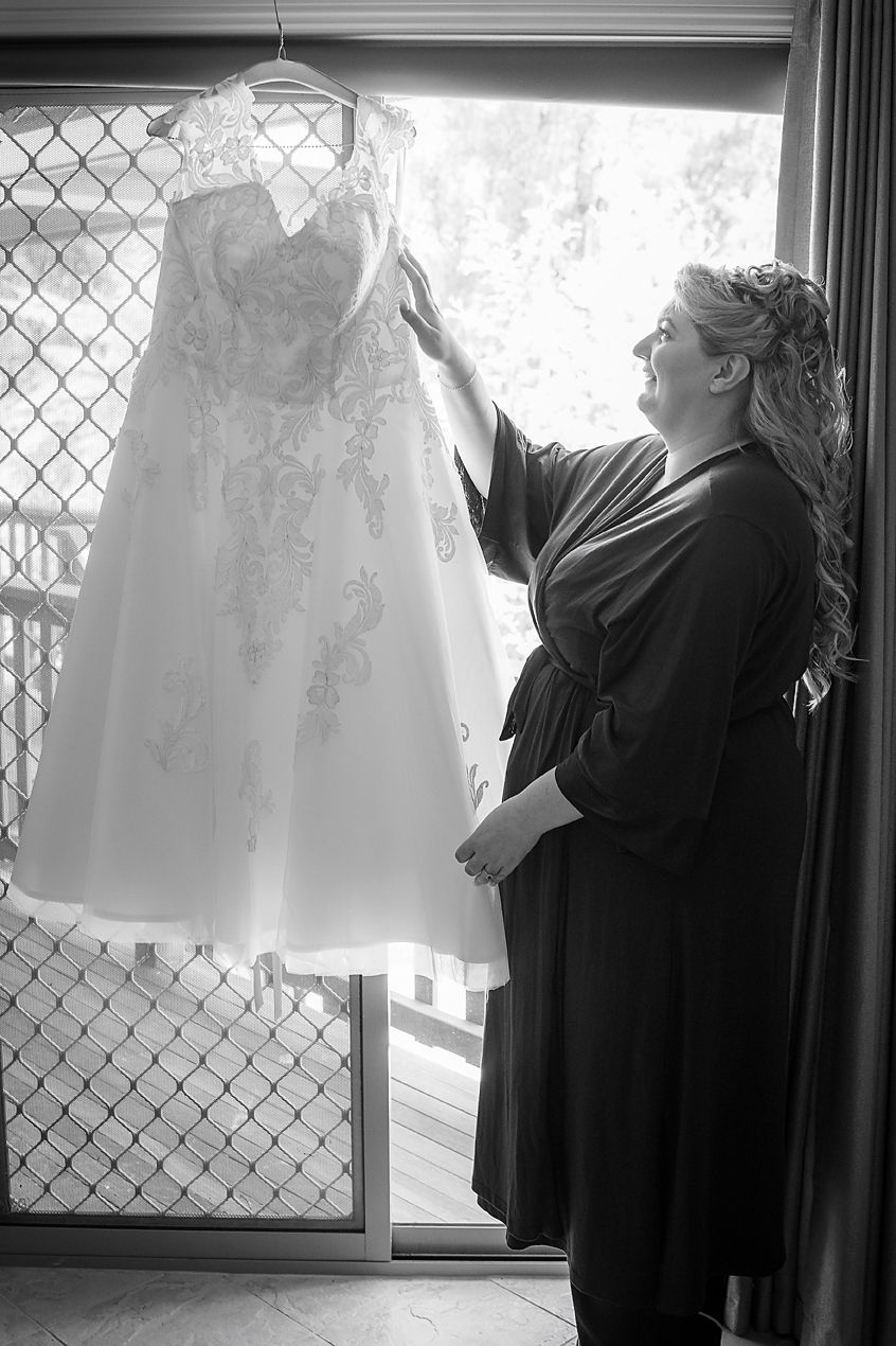 A person wearing a black robe adjusts a delicate white lace wedding dress hanging near a window.