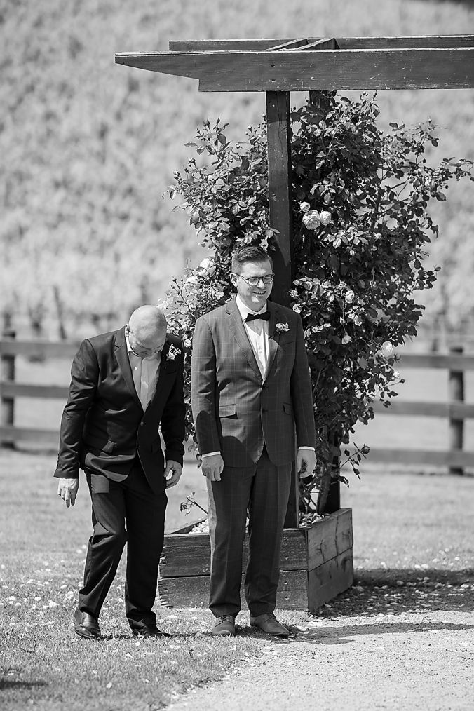 Two formally dressed people stand together under a wooden arch with decorative shrubs at an outdoor venue.