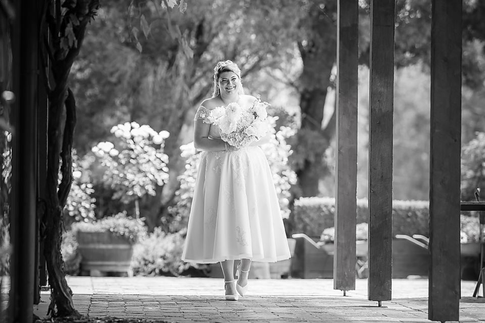 A black and white photo captures a bride holding a bouquet while standing between wooden pillars in a garden setting.