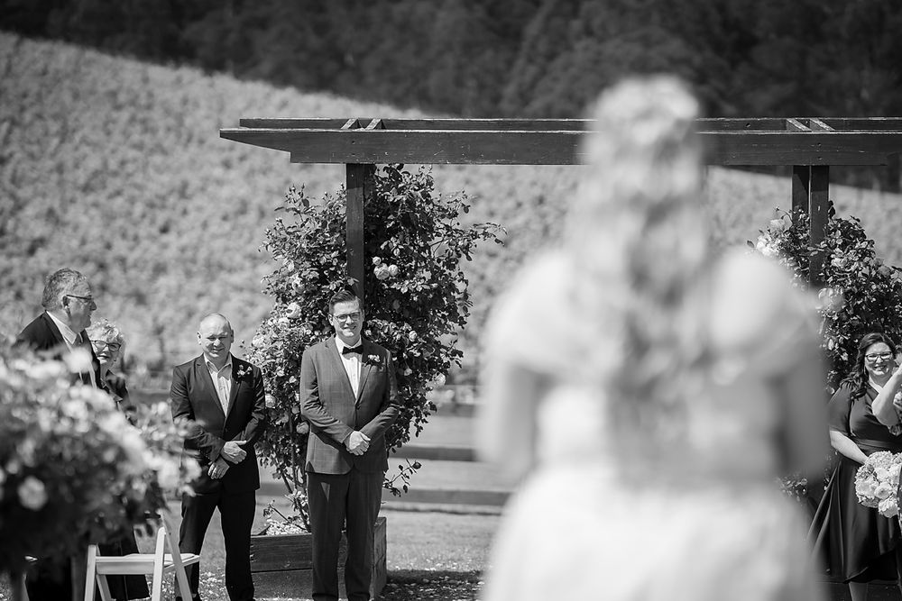Black and white outdoor wedding ceremony photo with out-of-focus bride in foreground and guests in background.