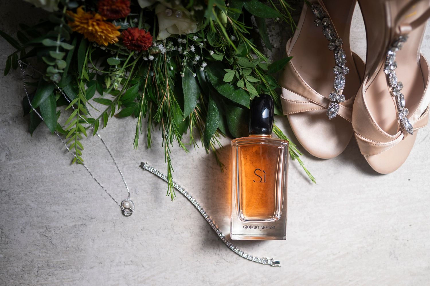Wedding accessories including blush satin heels, perfume bottle, and fresh greenery arranged on light gray surface.