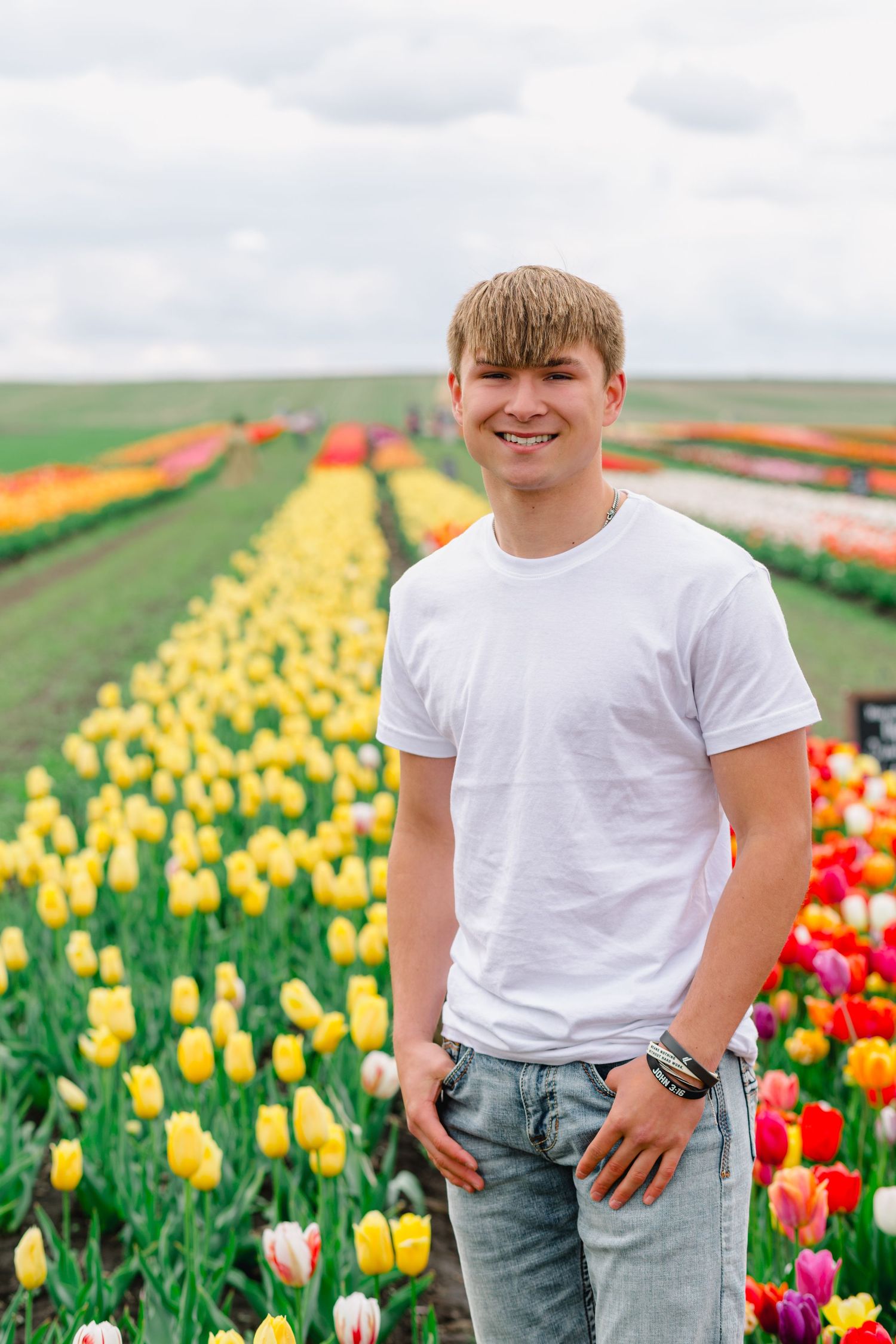 A child poses in a white t-shirt among vibrant rows of orange, yellow and red tulips in a blooming spring field.