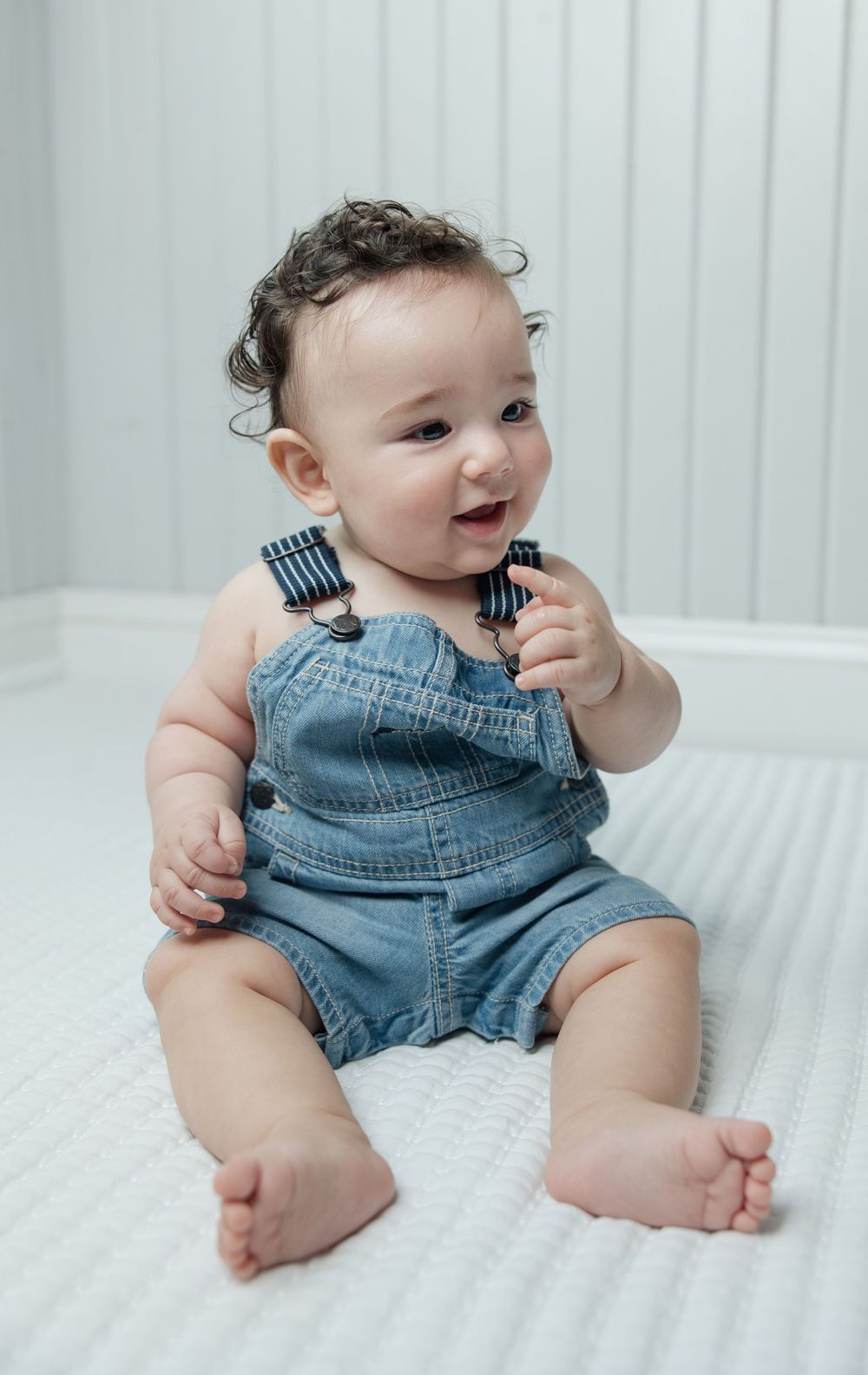 Smiling baby in jean overalls posing on white textured bedding.