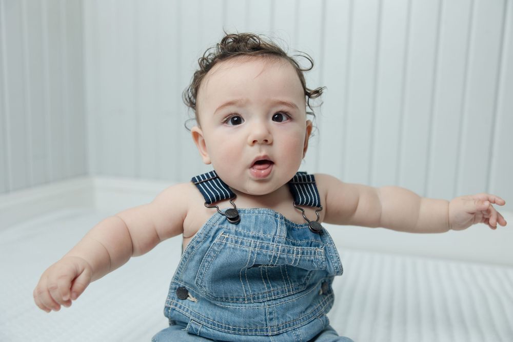 Baby wearing blue denim overalls lies on white textured bedding.