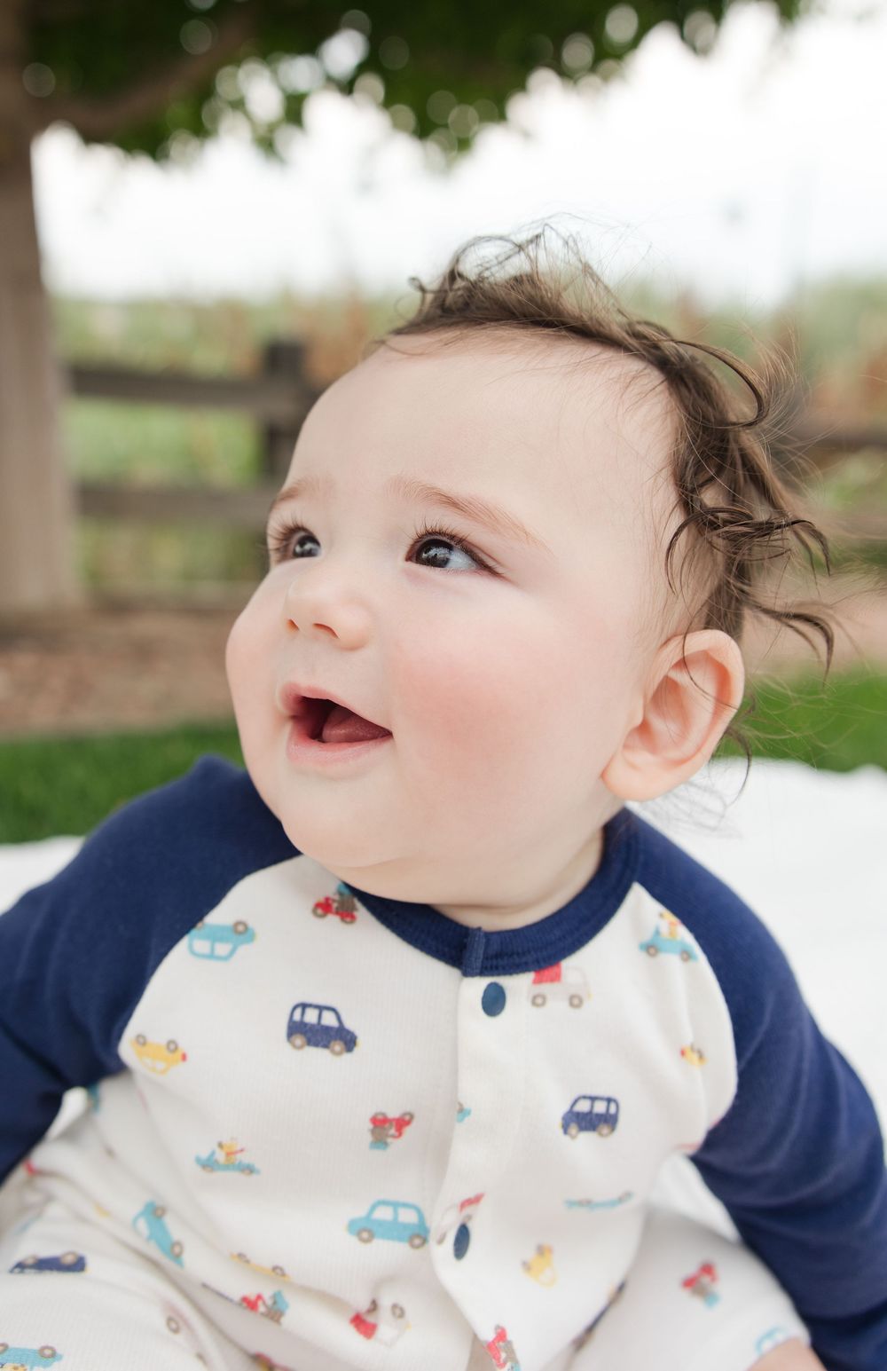 Baby wearing navy and white patterned outfit with car designs smiles outdoors.