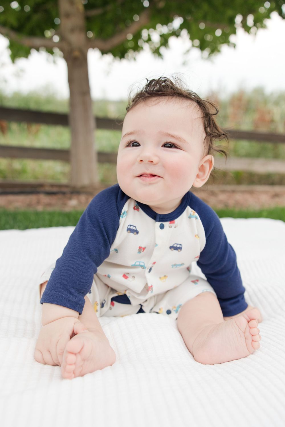 Baby in navy and white outfit lies on white blanket with trees in background.