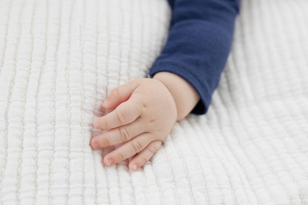 Infant wearing blue clothing reaches out while resting on white blanket.