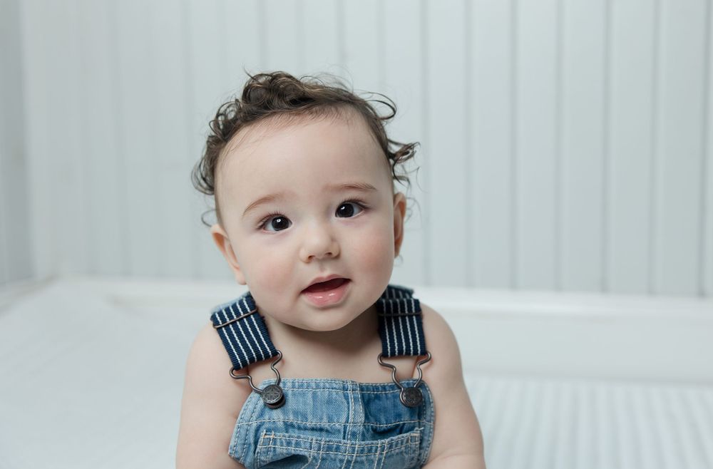 Adorable toddler in denim overalls sitting against a white background with a sweet expression.