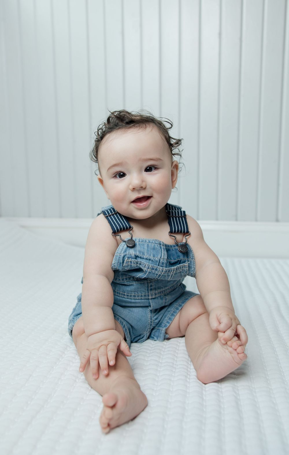 Baby wearing blue denim overalls sits on white bedding showing different playful expressions.