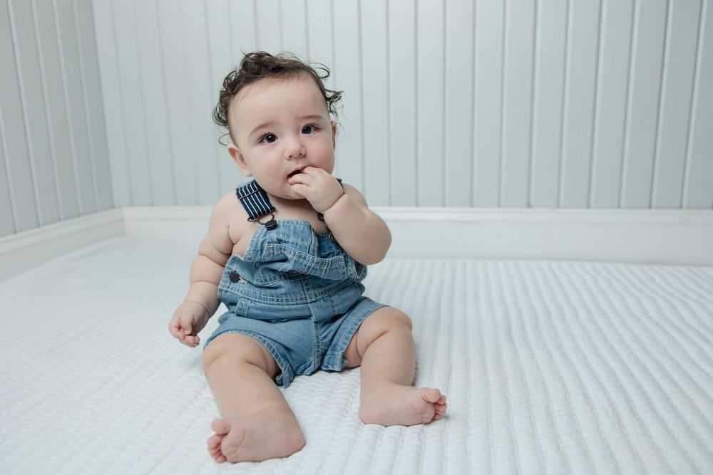 Adorable baby in denim overalls sitting on white bedding with thoughtful expression.