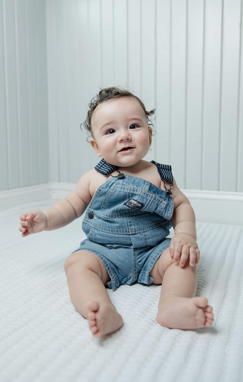 Baby wearing denim overalls sits on white surface with striped background.
