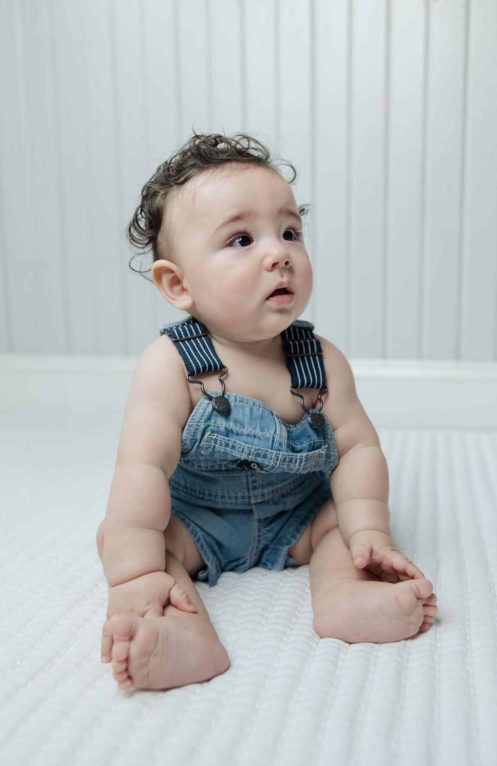 Baby in blue jean overalls sits against white backdrop looking to the side.