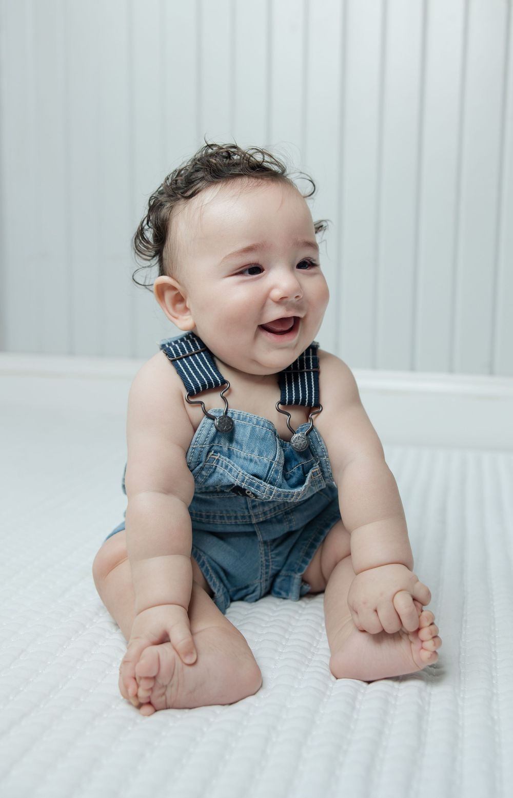 Happy baby wearing blue denim overalls sits on white textured surface.