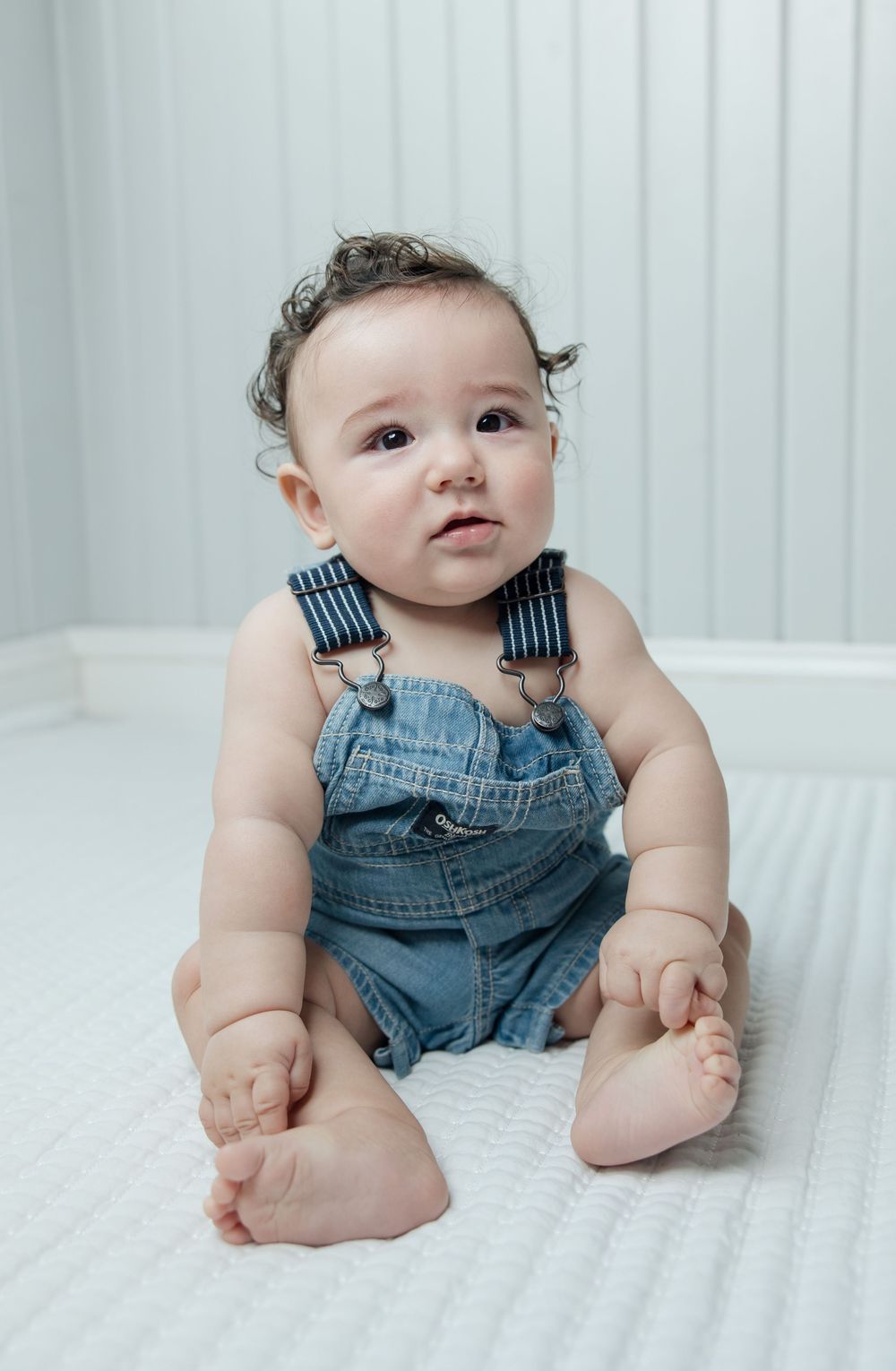Little one in blue jean overalls sitting on white surface with curious expression.