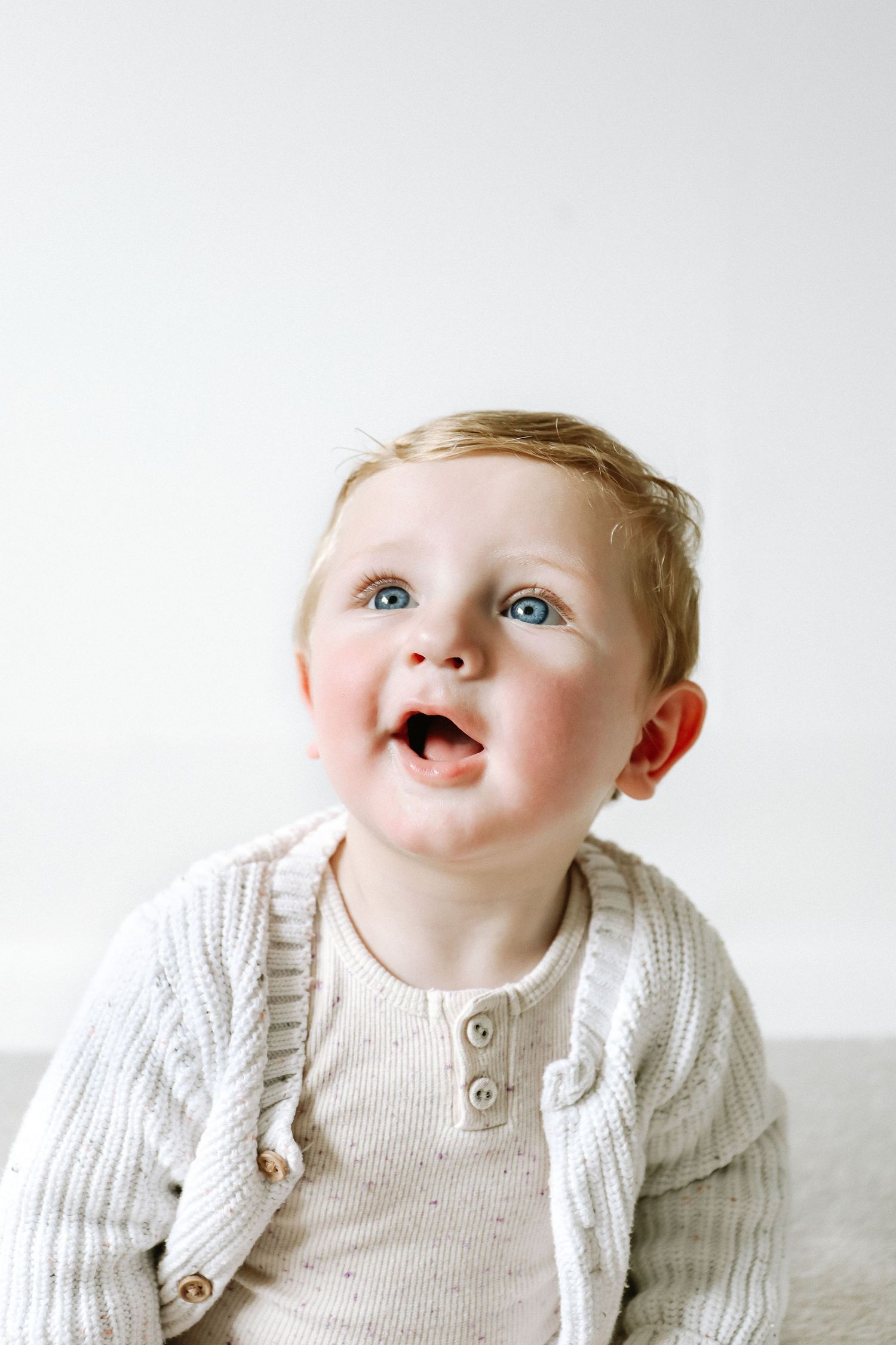 A joyful baby wearing a cozy white knit sweater cardigan looks up with an excited expression during birthday shoot in Nottingham