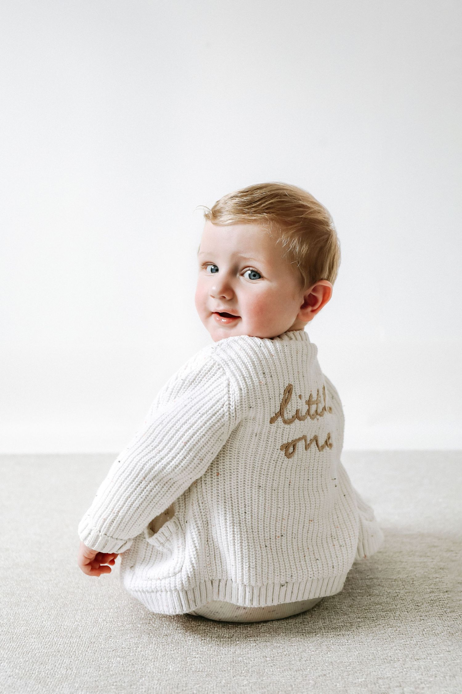 A baby wearing a white knit sweater smiles sitting on a light grey rug in front of white background, during birthday shoot in Nottingham