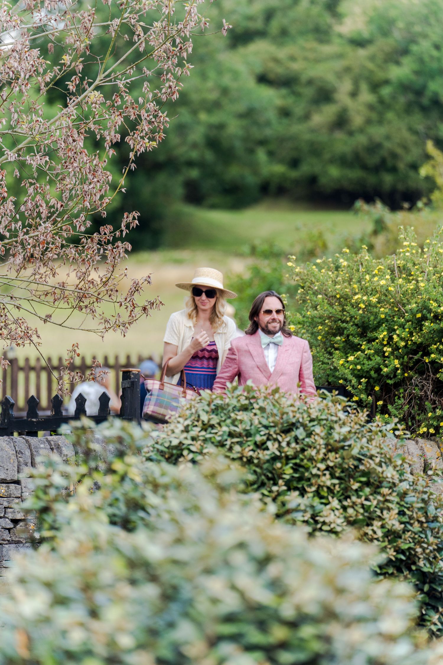 Two people walking along a garden path surrounded by lush greenery and flowering plants in summer bloom.