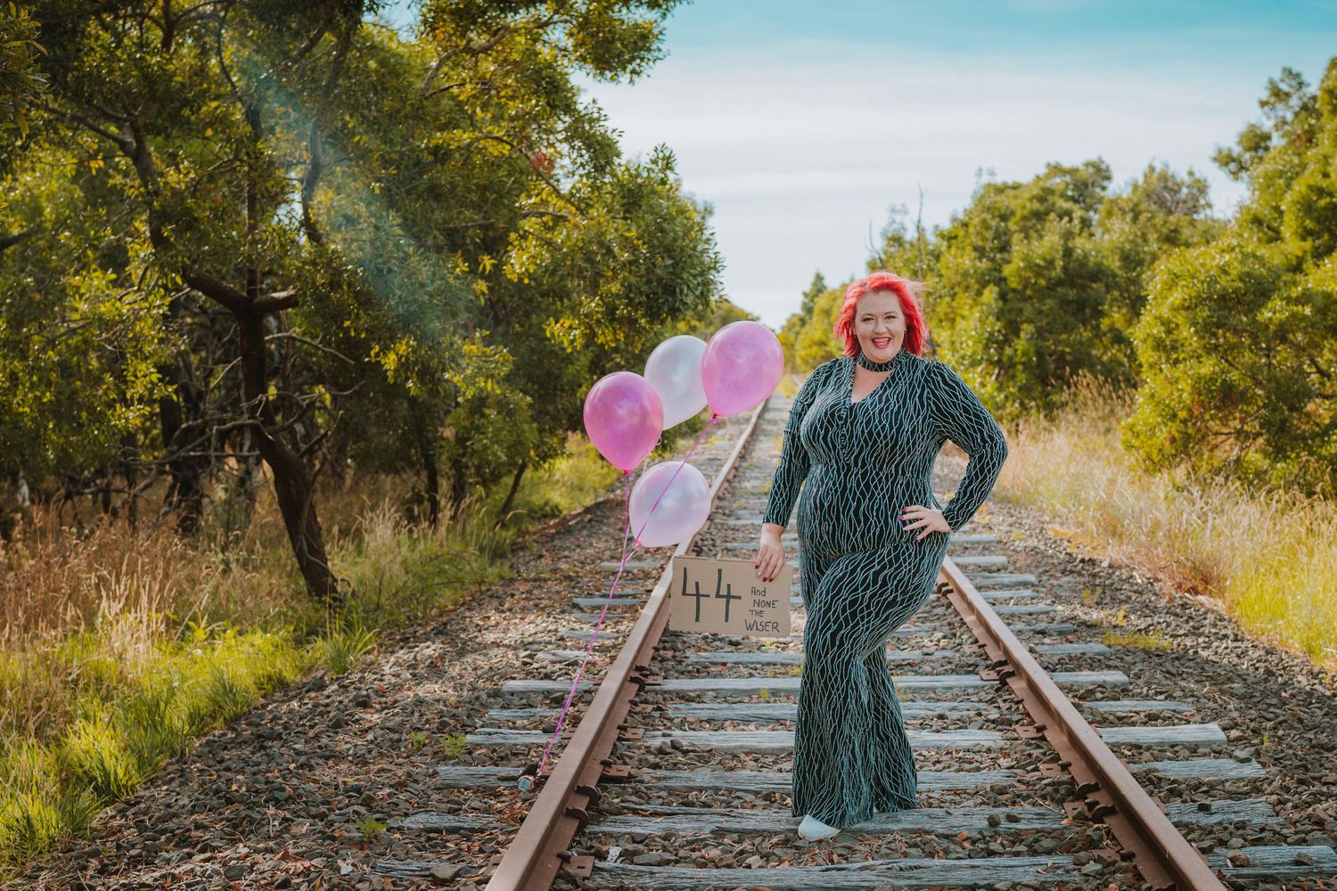 A figure in a green dress stands between railroad tracks holding pink balloons in rural scenery.