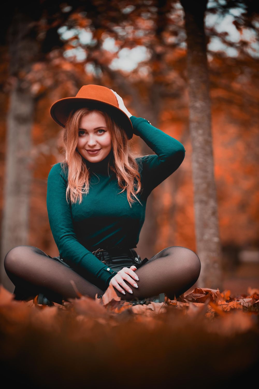 Autumn fashion photoshoot in a forest with emerald sweater and brown hat against orange fallen leaves.