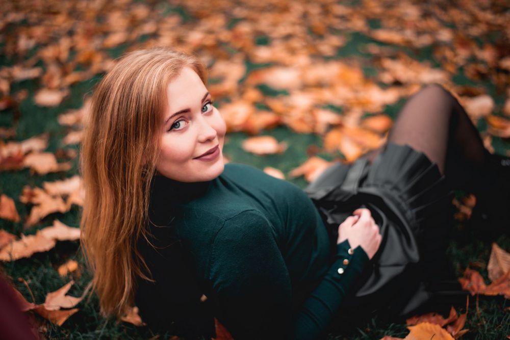 Someone in a dark green sweater lying on autumn leaves with long red hair.