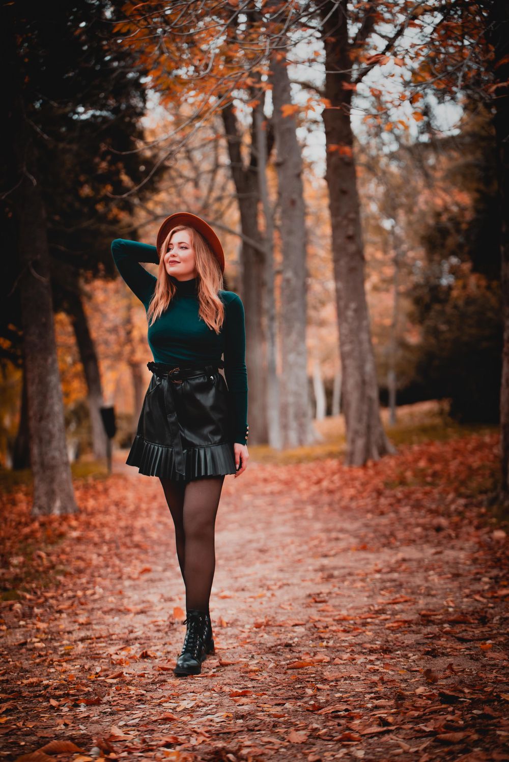 An autumn fashion portrait features a black pleated skirt and sweater outfit amid fallen orange leaves in a wooded path.