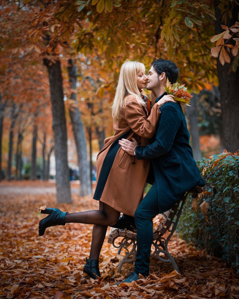 A romantic autumn scene with a couple embracing on a park bench surrounded by falling orange leaves and trees.