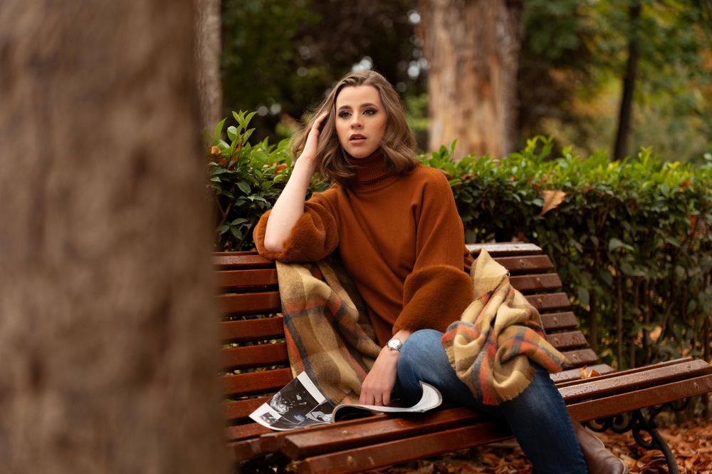 Someone in a brown sweater sitting on a wooden park bench surrounded by greenery.