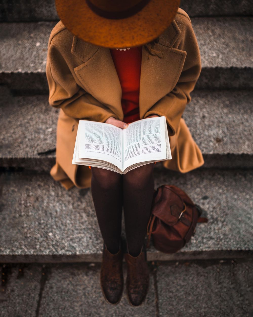 An overhead view shows someone in a brown coat reading a book while sitting on concrete steps with a leather bag nearby.
