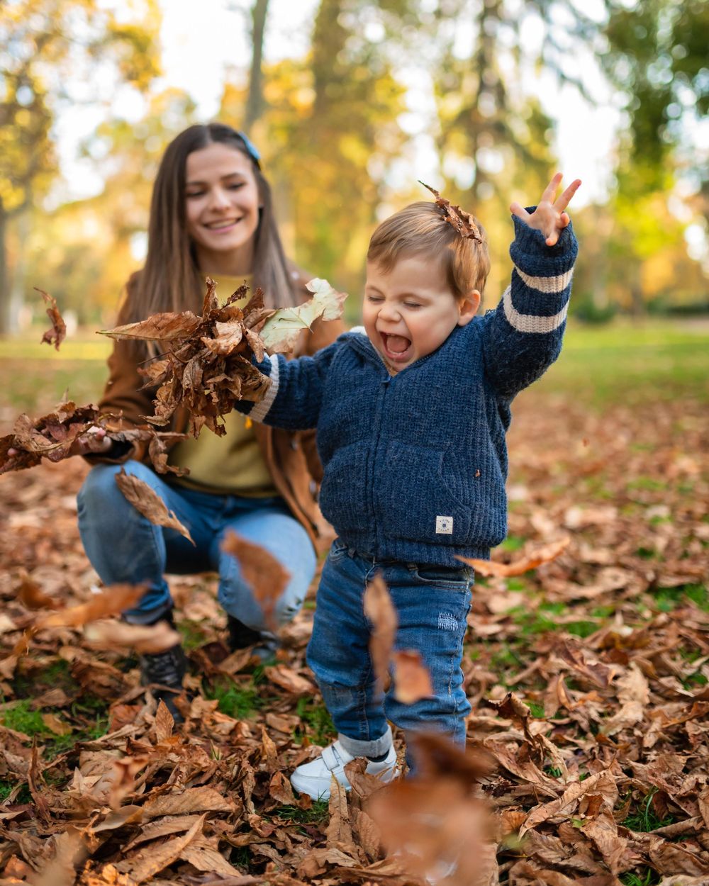 Joyful child playing in a pile of autumn leaves while an adult throws foliage in a sunny park setting.