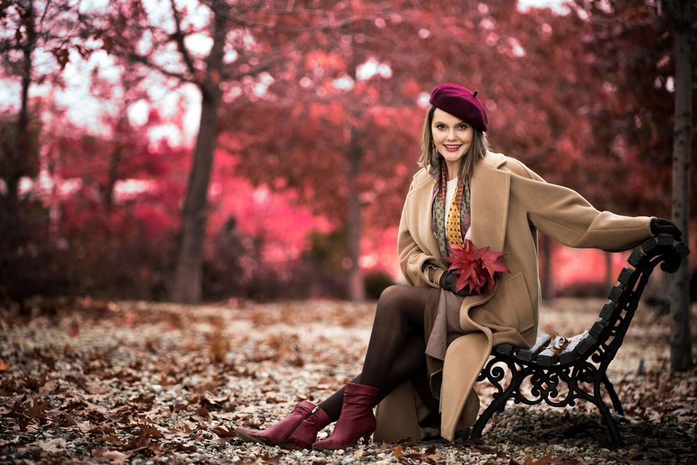 Person in tan coat and burgundy boots sits gracefully on park bench surrounded by pink autumn foliage.