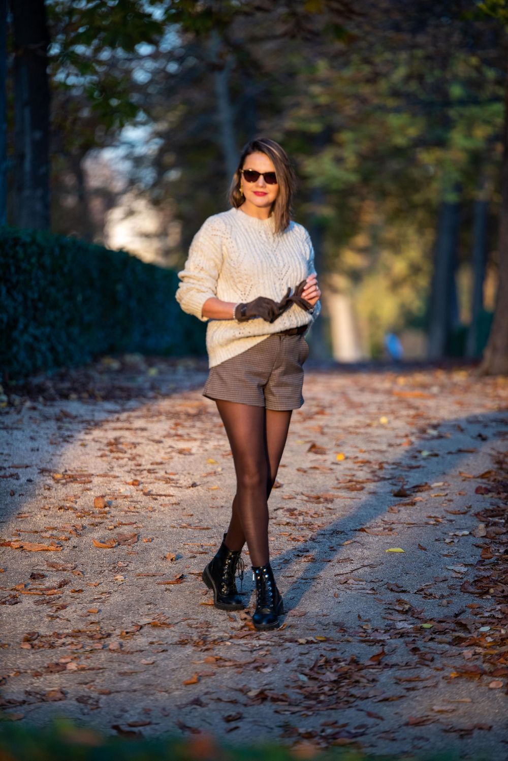 Lower body view of someone wearing a cream sweater and brown skirt walking through autumn leaves in a park.