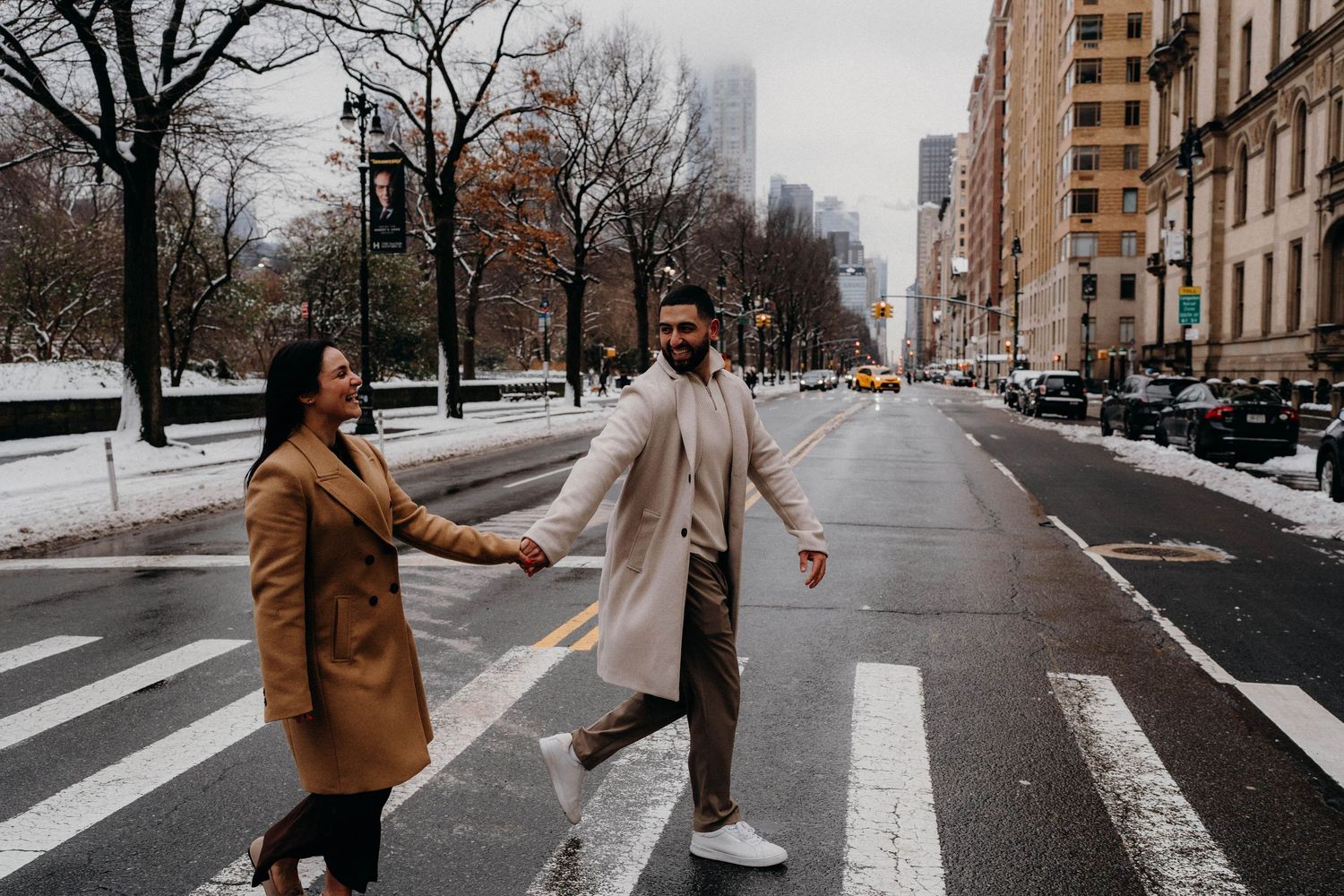 A playful couple walking across a snowy crosswalk in winter coats on a New York city street.