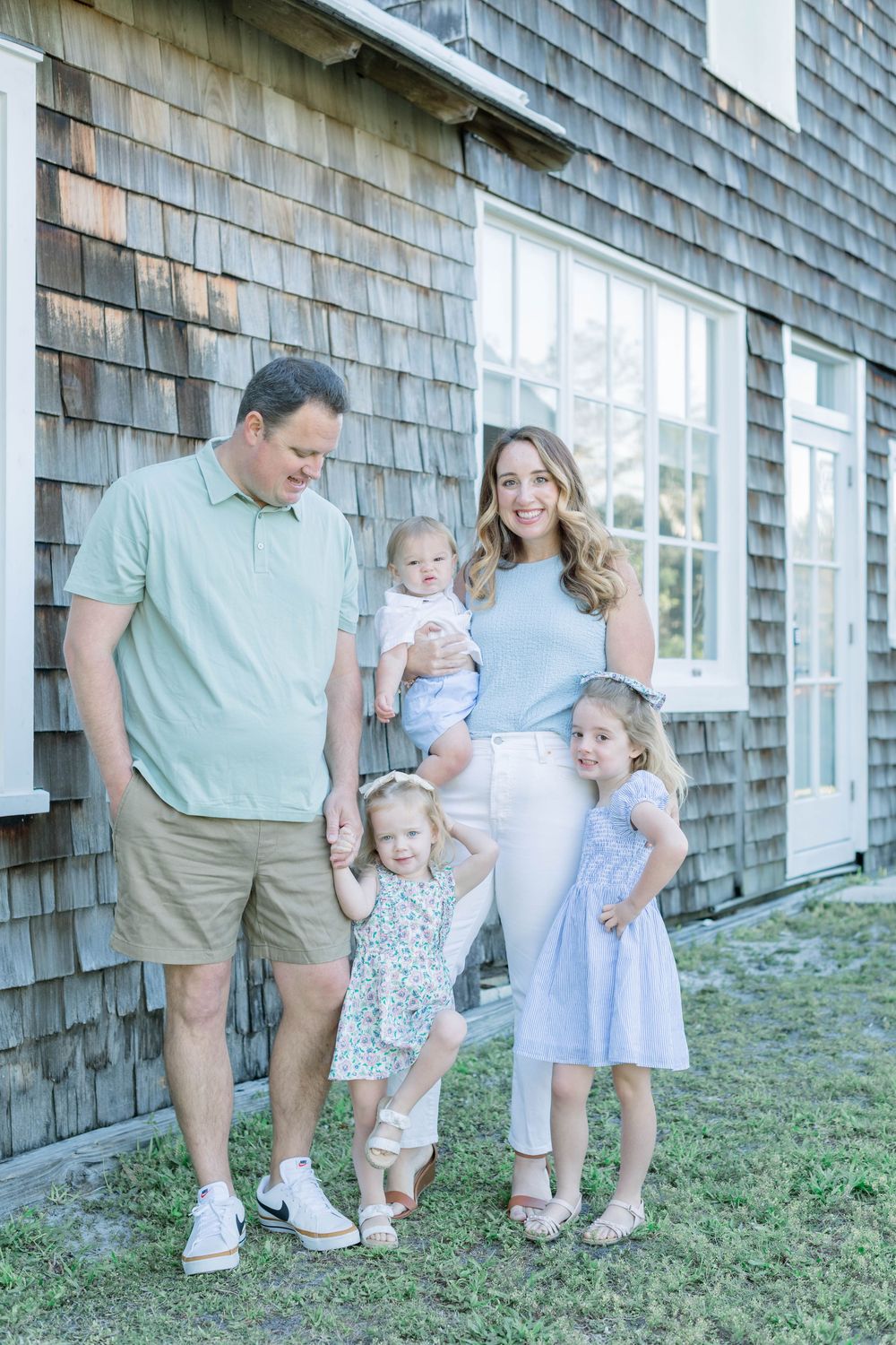Family dressed in light blue and neutral tones poses together against a rustic shingle-sided house on a sunny day.