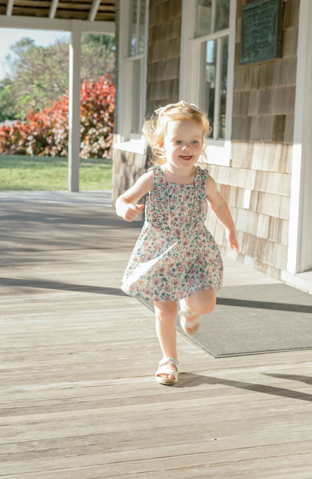 A young child in a floral sundress runs happily along a sunny covered porch on a summer day.