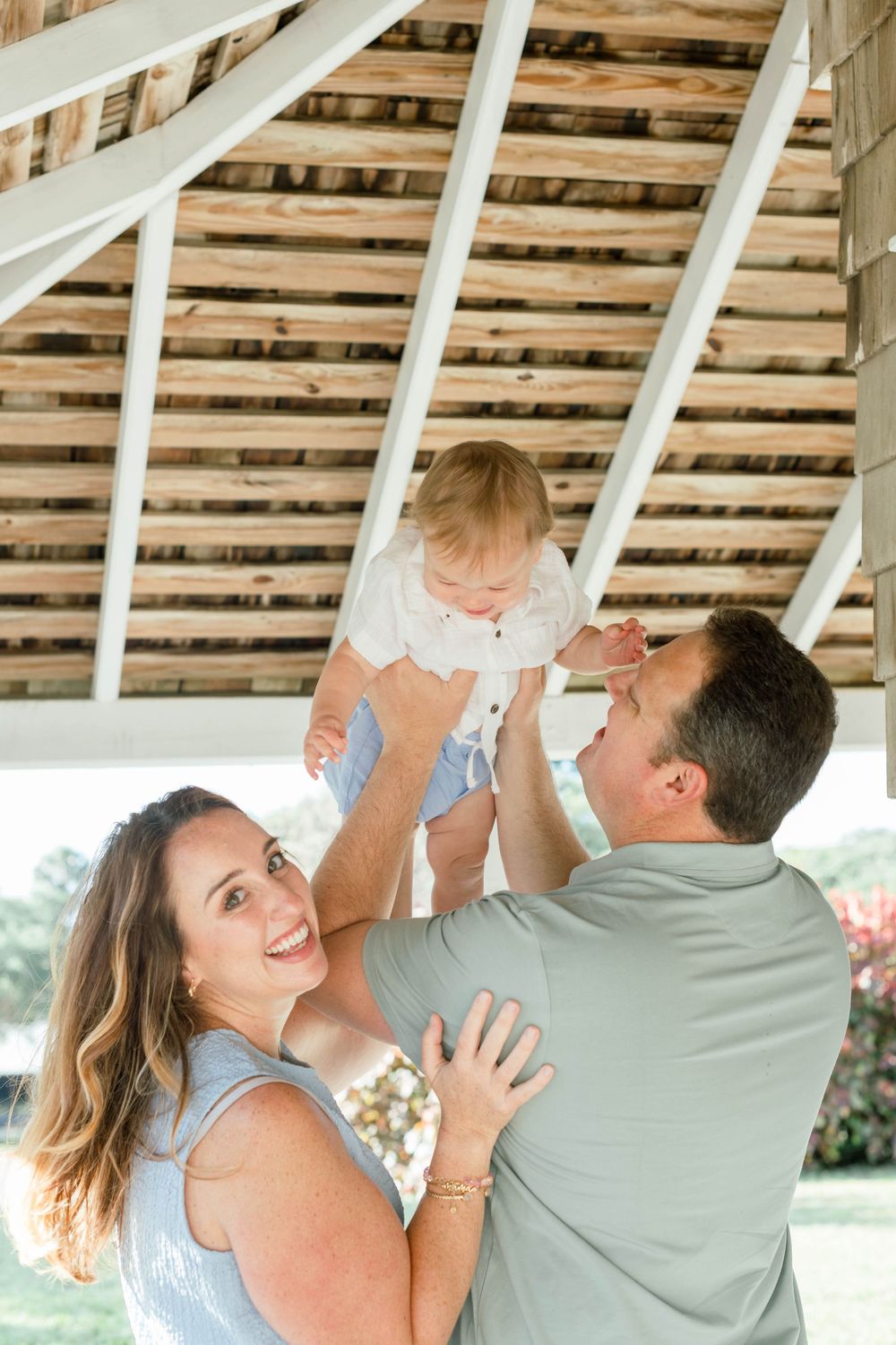 A joyful family moment captured under a wooden pergola as parents playfully lift their baby in the air.