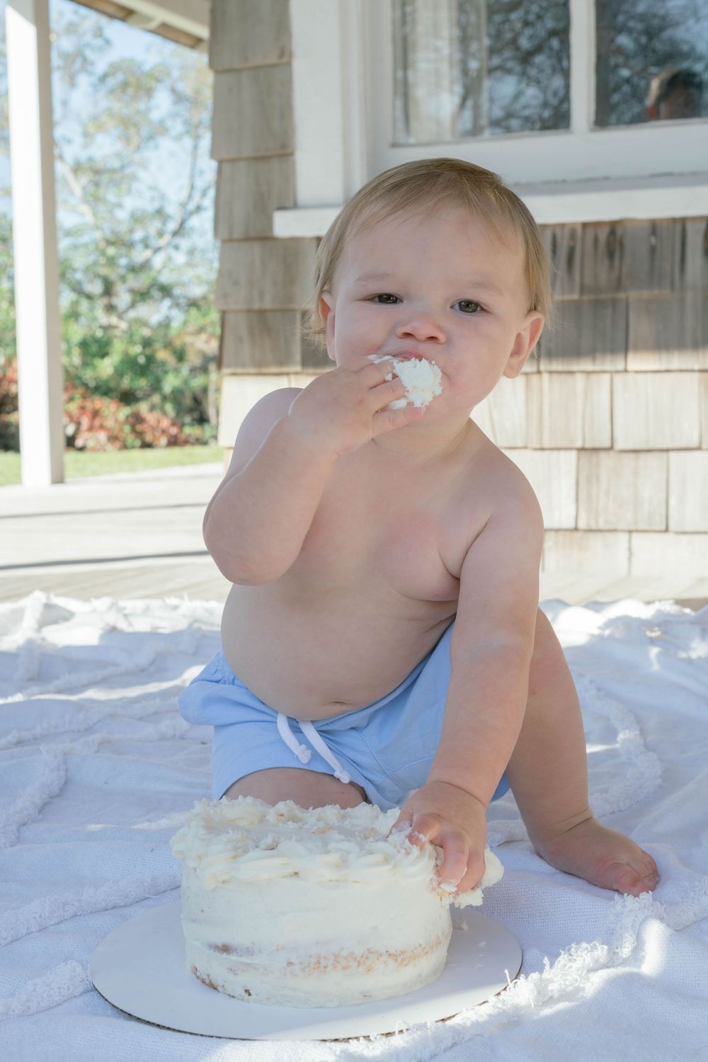 Small child sits on white blanket eating birthday cake with white frosting on outdoor porch.