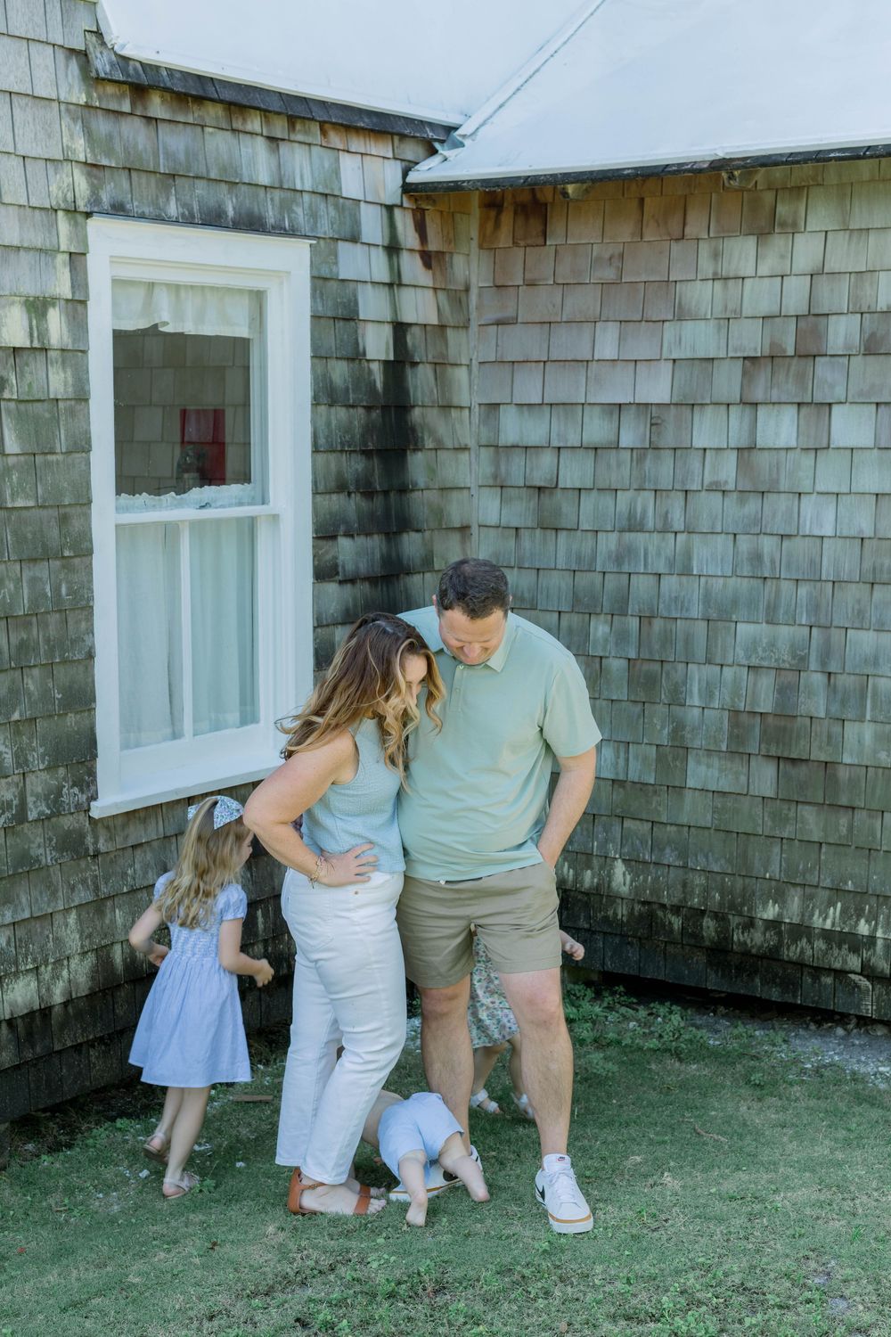 Family plays together outside a rustic gray shingle-sided house on the lawn in casual summer outfits.