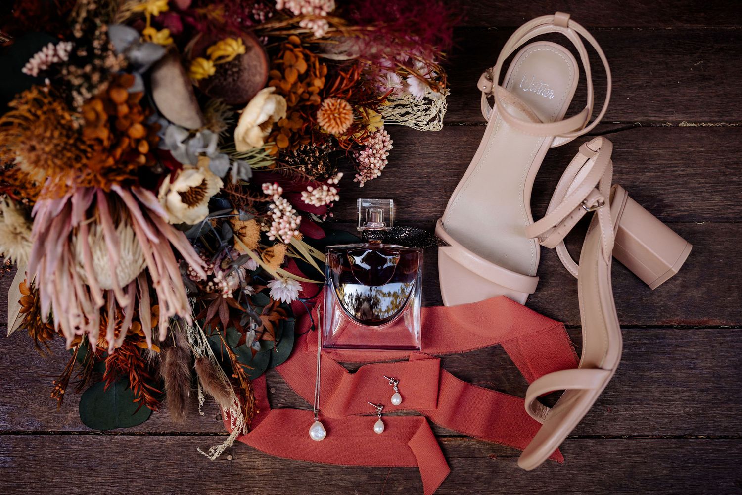 Wedding accessories with dried flowers, perfume bottle, nude heels and red envelope on dark wooden background.
