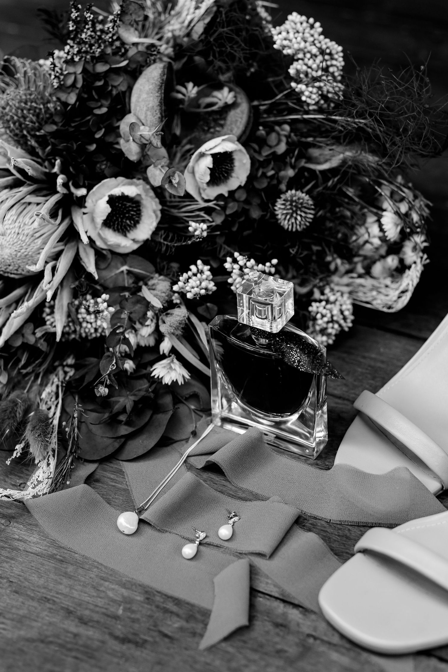 A black and white photo of a perfume bottle with a floral bouquet, earrings and ribbons on a wooden surface.