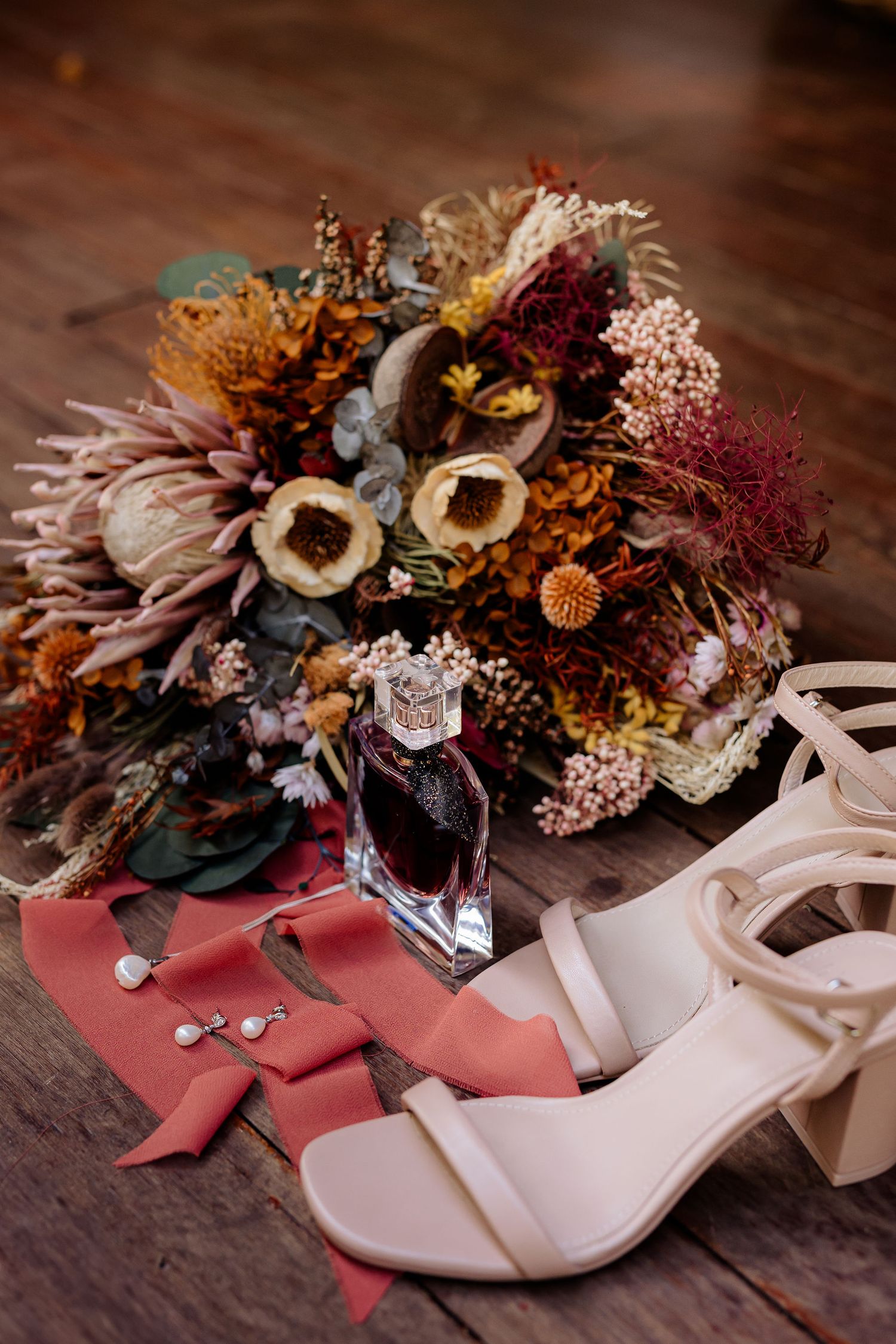 Wedding details with dried autumn floral bouquet, white block heel sandals and pink silk ribbon on wooden surface.