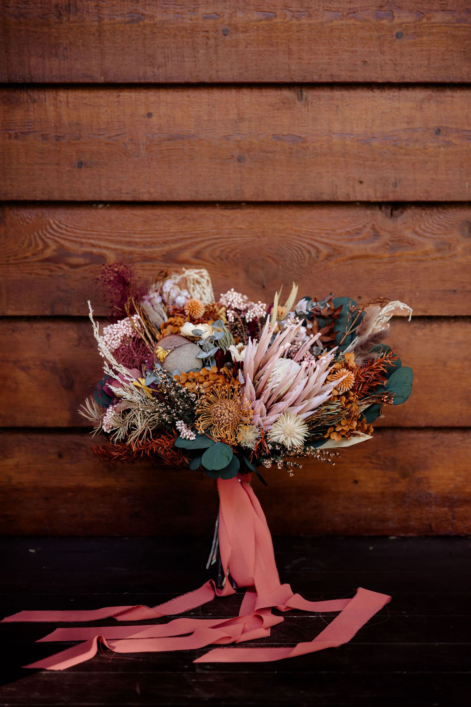 A rustic dried flower bouquet with pink protea, eucalyptus and burgundy blooms tied with a coral ribbon against wooden planks.
