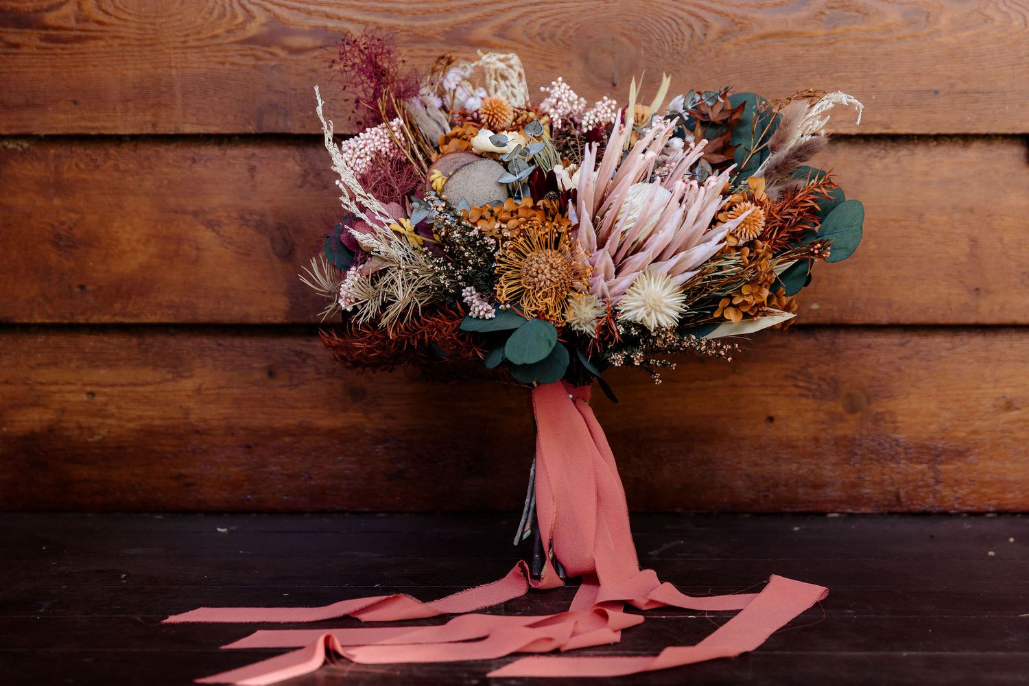 Rustic wedding bouquet with pink protea, dried flowers, eucalyptus and coral silk ribbon on wooden background.