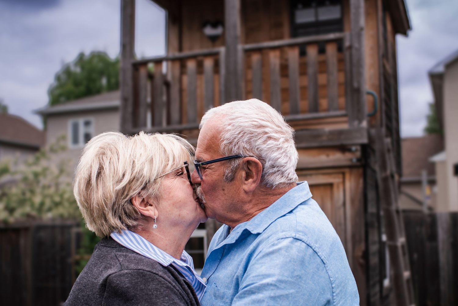 Two seniors share an intimate embrace and kiss outdoors against a gentle blurred background.