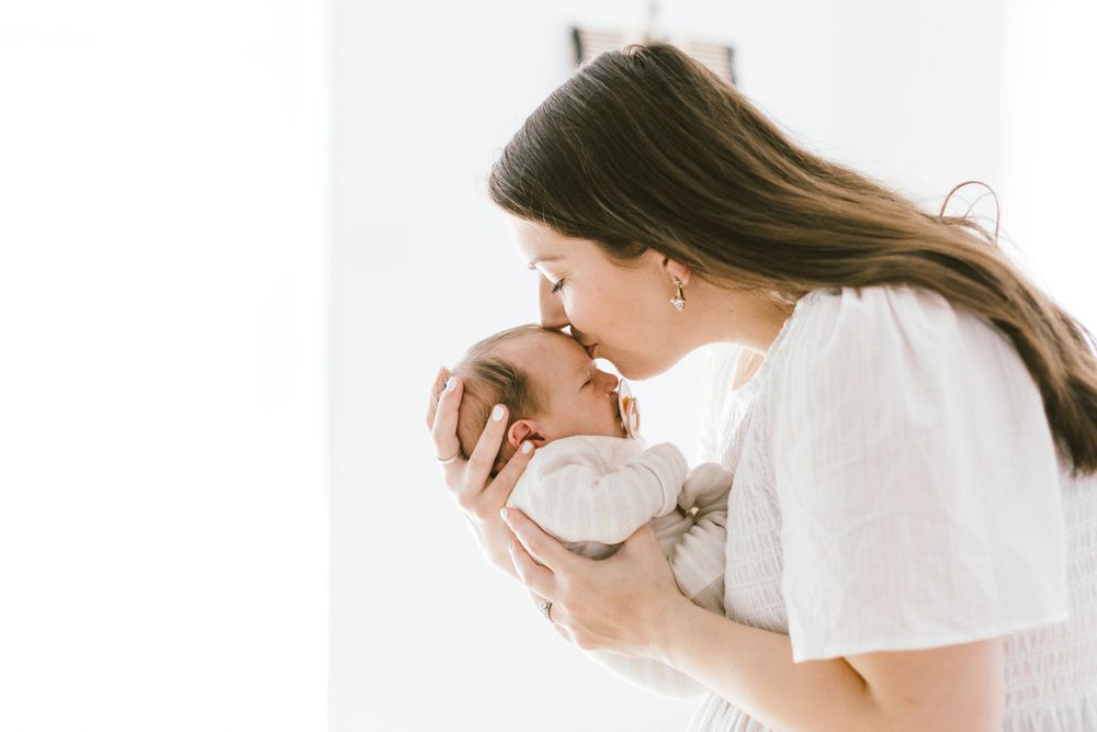 Mother in white giving gentle kiss to newborn baby in bright room setting.