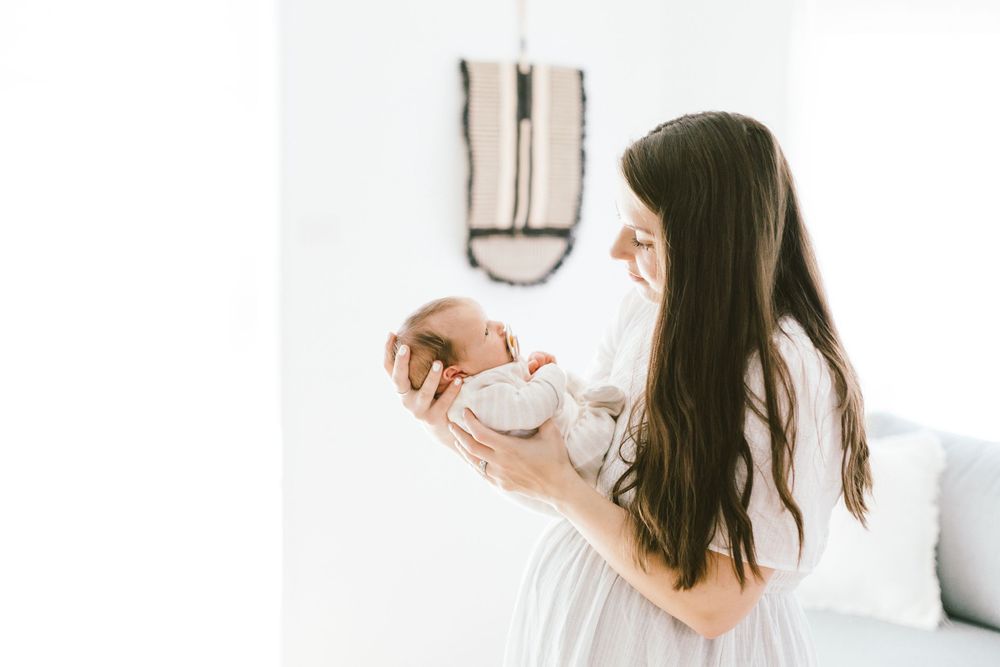 Woman in white dress gazes lovingly at newborn baby against minimalist background.