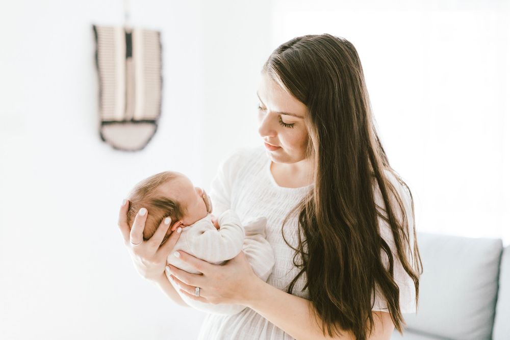 Woman in white dress smiles while holding sleeping newborn in bright minimal room.