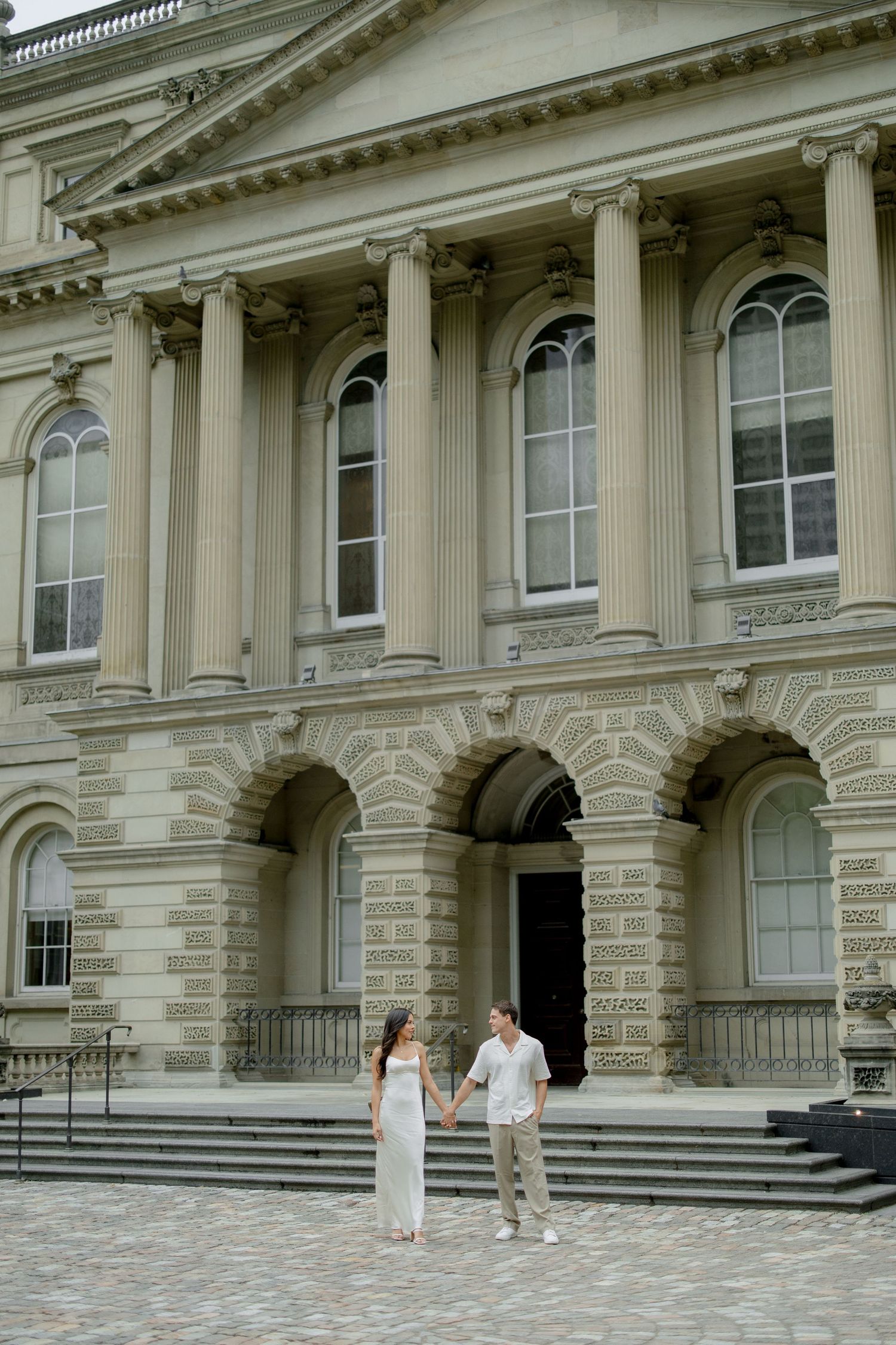 A couple poses in front of a grand neoclassical building with elegant columns and stone architecture.
