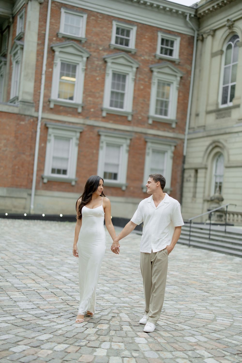 A romantic couple walks hand in hand on cobblestone pavement outside a historic brick building in formal attire.