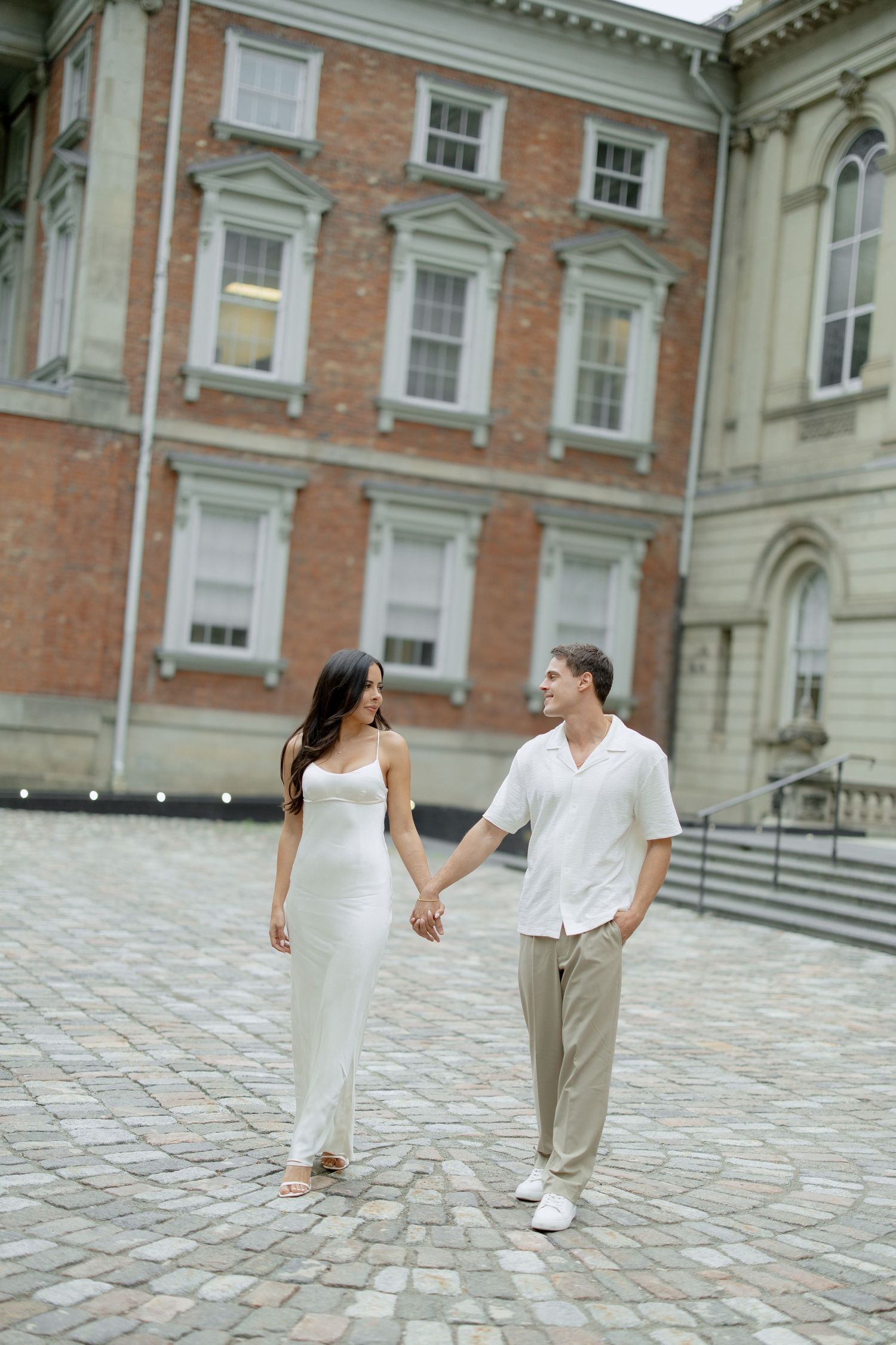 A romantic couple walks hand in hand on cobblestone pavement outside a historic brick building in formal attire.